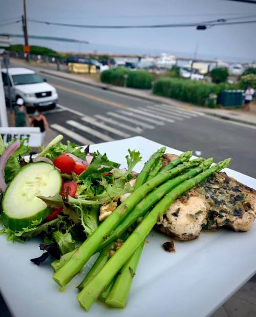 A plate of grilled chicken with asparagus and a fresh salad, set against a coastal street view.