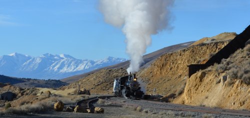 A steam locomotive chugs along a track with mountains in the background, releasing a plume of white smoke.