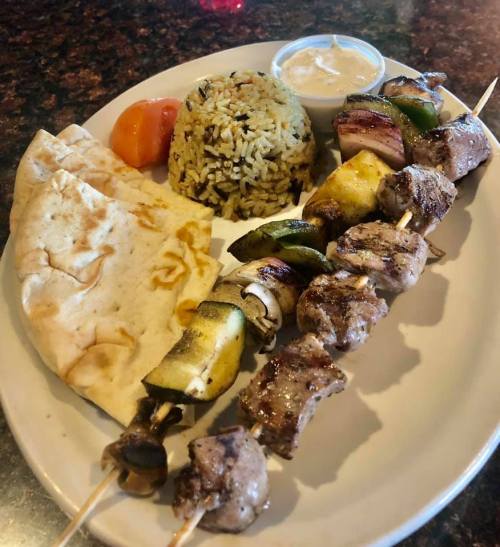 A plate of skewered meat and vegetables, served with rice, pita bread, and a small cup of sauce.