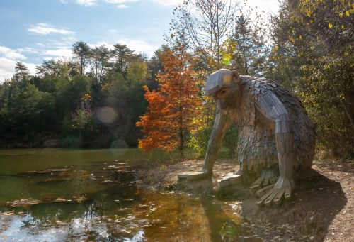 A large wooden sculpture of a creature sits by a serene pond, surrounded by trees and autumn foliage.