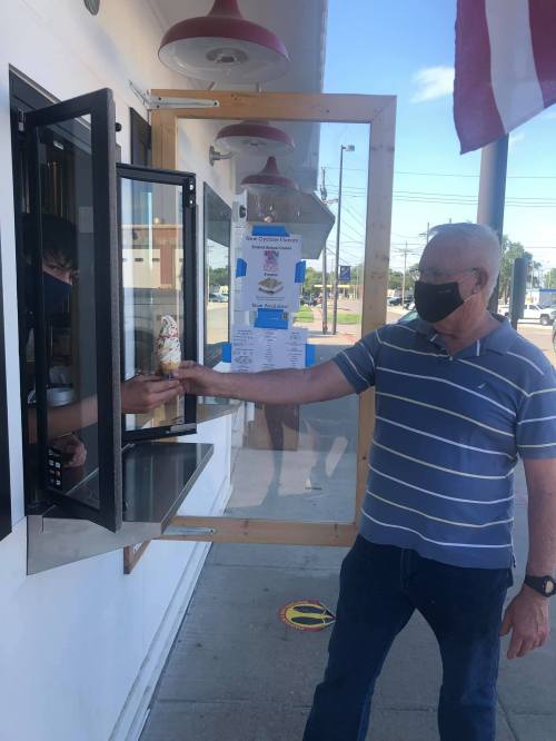 A man in a mask receives an ice cream cone from a server at a takeout window on a sunny day.