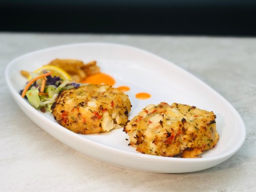 Two crab cakes on a white plate, accompanied by a side salad and drizzles of sauce.