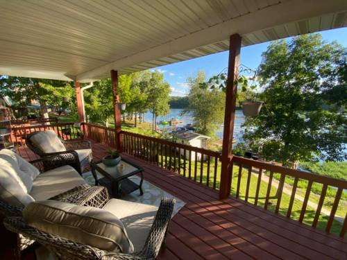 A cozy porch with wicker chairs overlooks a serene lake surrounded by trees and a dock in the distance.