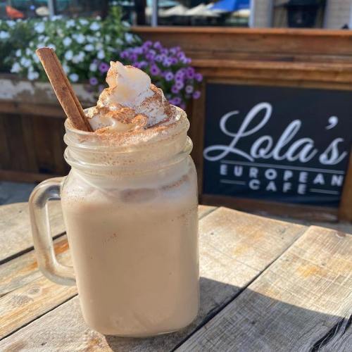 A frothy drink topped with whipped cream and cinnamon, served in a mason jar on a wooden table outside a café.