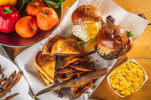 A wooden table with burgers, fried egg, sweet potato fries, mac and cheese, and fresh vegetables in bowls.