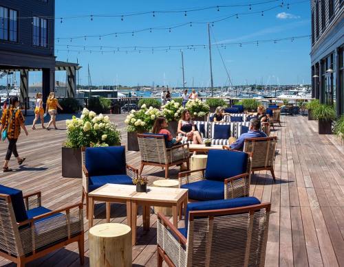 A sunny outdoor deck with seating, people walking, and boats in the background, surrounded by greenery and string lights.