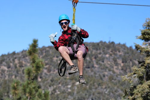 A person in a helmet and sunglasses zip-lining, giving a thumbs-up, with mountains in the background.