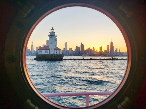 View of a lighthouse and city skyline at sunset, framed by a circular window on a boat.