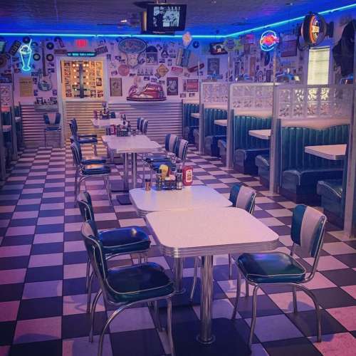 A retro diner interior with checkered floors, teal booths, and vintage decor, featuring empty tables and neon signs.