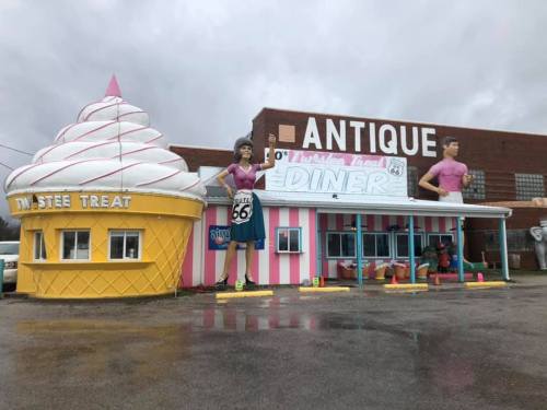 Colorful diner with giant ice cream cone and statues of two people, featuring a retro Route 66 theme. Overcast sky.