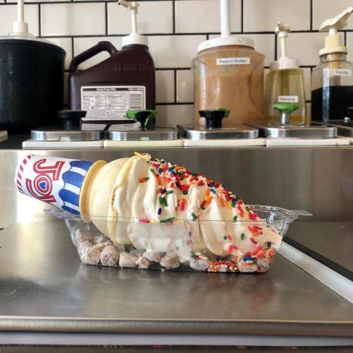 A soft serve ice cream cone topped with sprinkles, served in a dish with crushed toppings.