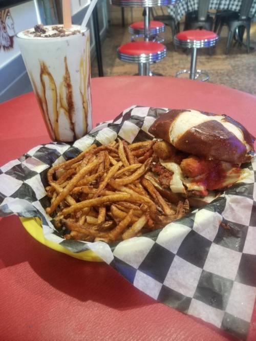 A plate with a pretzel bun sandwich, fries, and a tall milkshake on a red table in a diner setting.