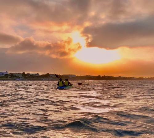 Two people kayak on a shimmering water surface at sunset, with a colorful sky and distant shoreline in the background.
