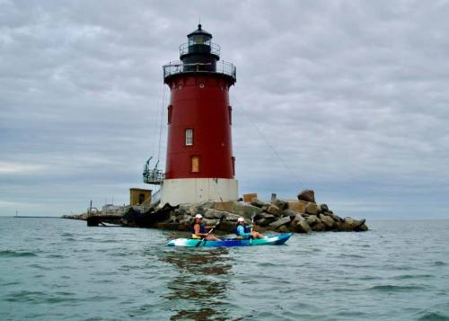 Two kayakers paddle near a red lighthouse on a rocky shore under a cloudy sky.