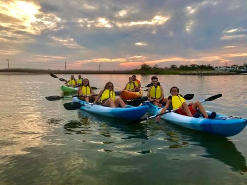 A group of people in colorful kayaks paddling on calm water during sunset, wearing life jackets.