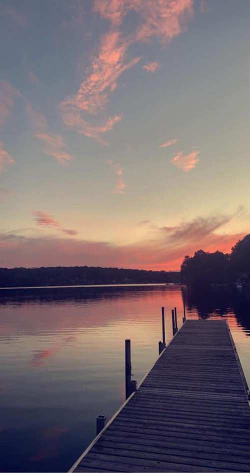 A serene lake at sunset, with a wooden dock extending into calm waters reflecting pink and orange hues in the sky.