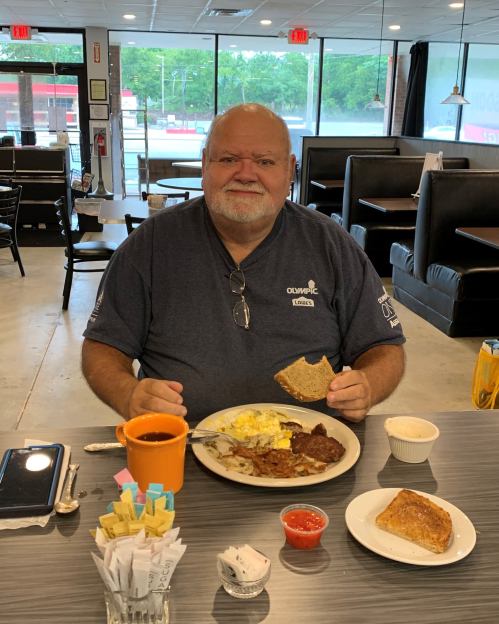 A man sits at a table in a diner, smiling with a plate of breakfast food, coffee, and toast in front of him.