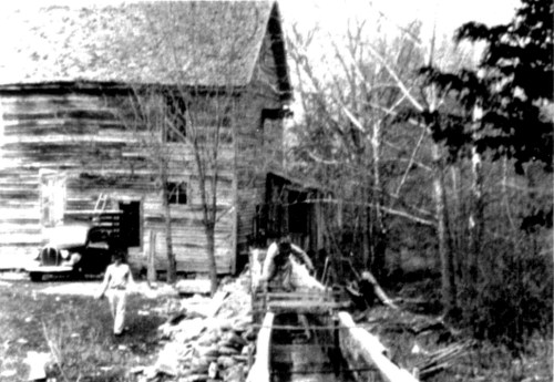 A black-and-white photo of a rustic wooden building, a small bridge, and a person walking near a stream.