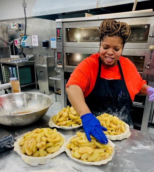 A woman in an orange shirt and gloves prepares apple pies in a kitchen, placing apple filling into pie crusts.
