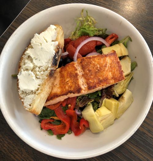A bowl with grilled salmon, mixed greens, red peppers, artichokes, and a slice of bread with cream cheese.