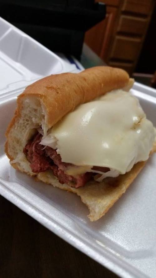 A close-up of a sandwich with layers of meat and melted cheese in a soft bread roll, served in a takeout container.