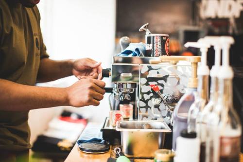 A person adjusts a coffee machine on a countertop, surrounded by various bottles and tools.