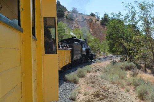 A vintage steam train curves around a hillside, viewed from a yellow train car window. Green trees and dry terrain surround it.