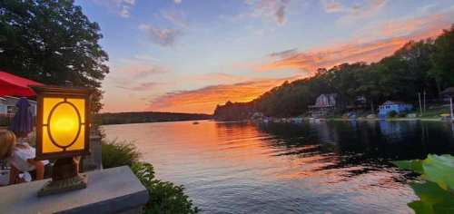 A serene lake at sunset, with vibrant orange and purple skies reflecting on the water, surrounded by trees and houses.