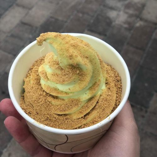 A hand holding a cup of green soft serve ice cream topped with a layer of crushed cookies or graham cracker crumbs.