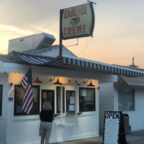 A person stands at the window of a vintage ice cream shop, "Dari Creme," with an American flag and a sign that says "Open."