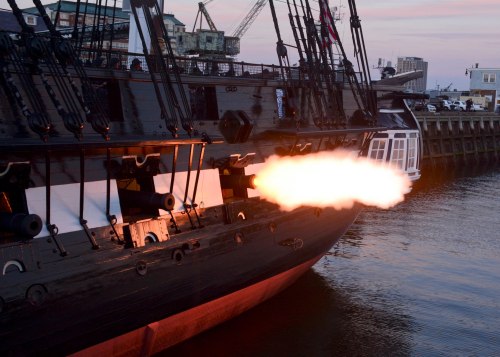 A historic ship fires a cannon, with flames and smoke visible, against a waterfront backdrop at sunset.