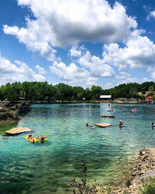 A serene lake with people swimming and floating on colorful rafts, surrounded by lush greenery and fluffy clouds.