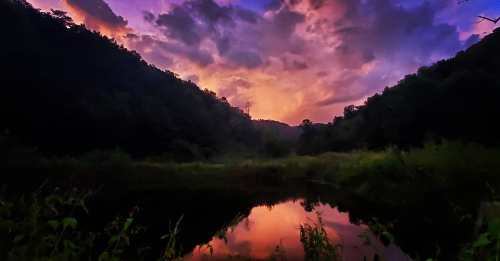 A serene landscape at dusk, featuring a calm pond reflecting vibrant purple and orange clouds between lush hills.