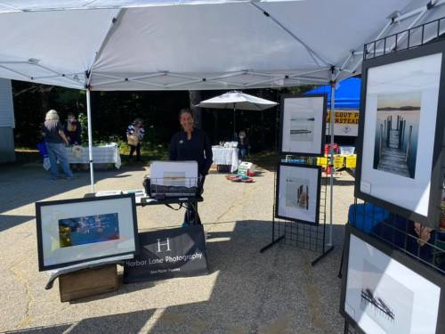 A vendor stands under a tent displaying framed photographs at an outdoor market.