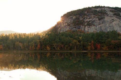 Sunrise over a mountain reflecting in a calm lake, surrounded by colorful autumn trees.