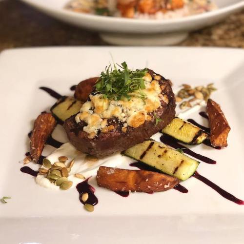 A gourmet dish featuring a grilled steak topped with cheese, served with roasted vegetables and a drizzle of sauce.