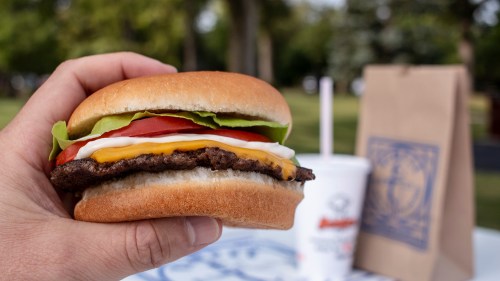 A hand holding a juicy burger with lettuce, tomato, cheese, and mayonnaise, with a drink and bag in the background.