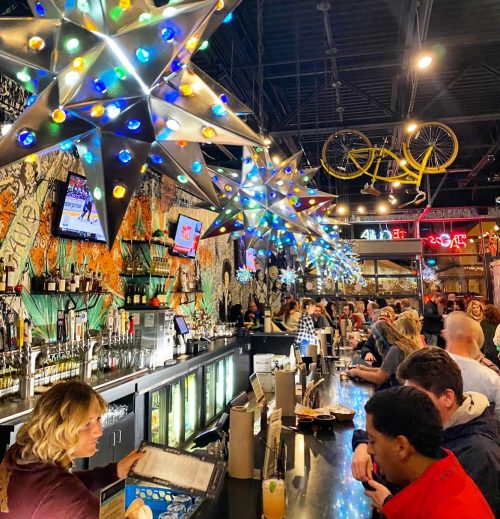 A lively bar scene with colorful star-shaped lights, patrons at the counter, and vibrant wall art in the background.