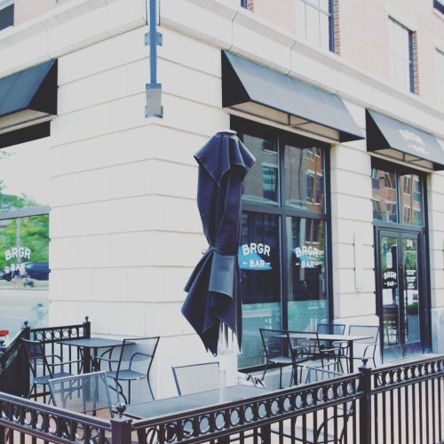 Exterior of BRGR Bar with a black umbrella, outdoor seating, and large windows on a sunny day.