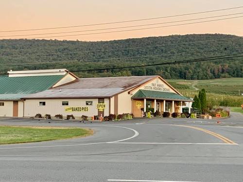 A roadside fruit market with a green roof, signs for baked pies, and a scenic mountain backdrop at sunset.