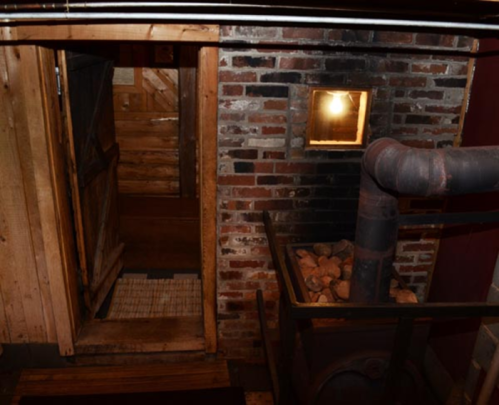 A rustic interior with a wooden door, brick walls, and a metal stove with rocks beside it, illuminated by a small window.