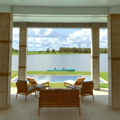A serene lakeside view from a porch with wooden chairs, a pool, and kayakers in the distance under a cloudy sky.