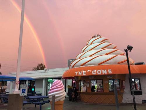 An ice cream stand with a whimsical cone-shaped roof under a colorful sky with two rainbows.
