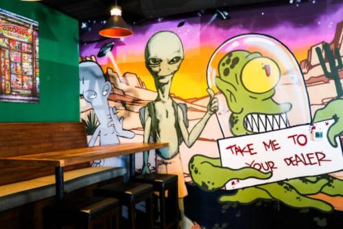 A colorful mural featuring aliens holding a sign that says "Take me to your dealer" in a trendy cafe setting.