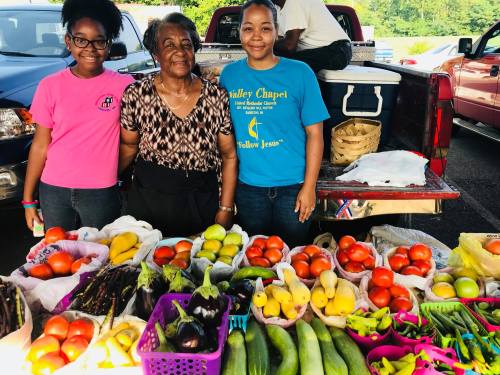 Three women stand behind a colorful display of fresh fruits and vegetables at a market, smiling at the camera.