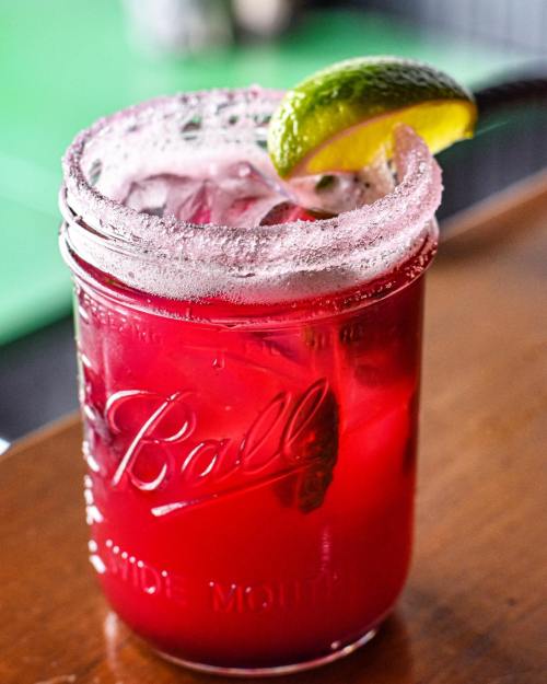 A vibrant red drink in a mason jar, garnished with a lime wedge and rimmed with salt.