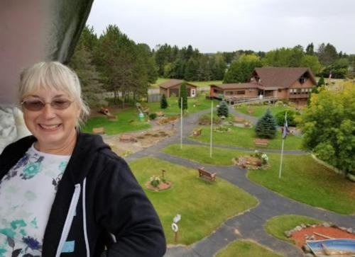 A smiling woman stands in front of a scenic view of a park with trees, pathways, and a large building in the background.
