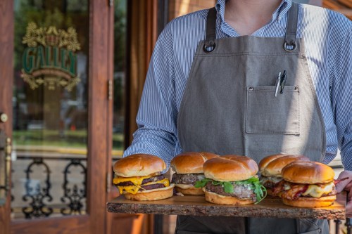 A person in an apron holds a wooden tray with five gourmet burgers outside a restaurant.
