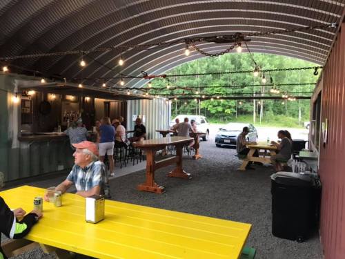 A lively outdoor bar with patrons enjoying drinks under string lights, surrounded by greenery and a rustic setting.