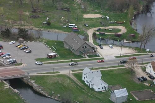 Aerial view of a park area with a building, parking lot, river, and several vehicles along a road.